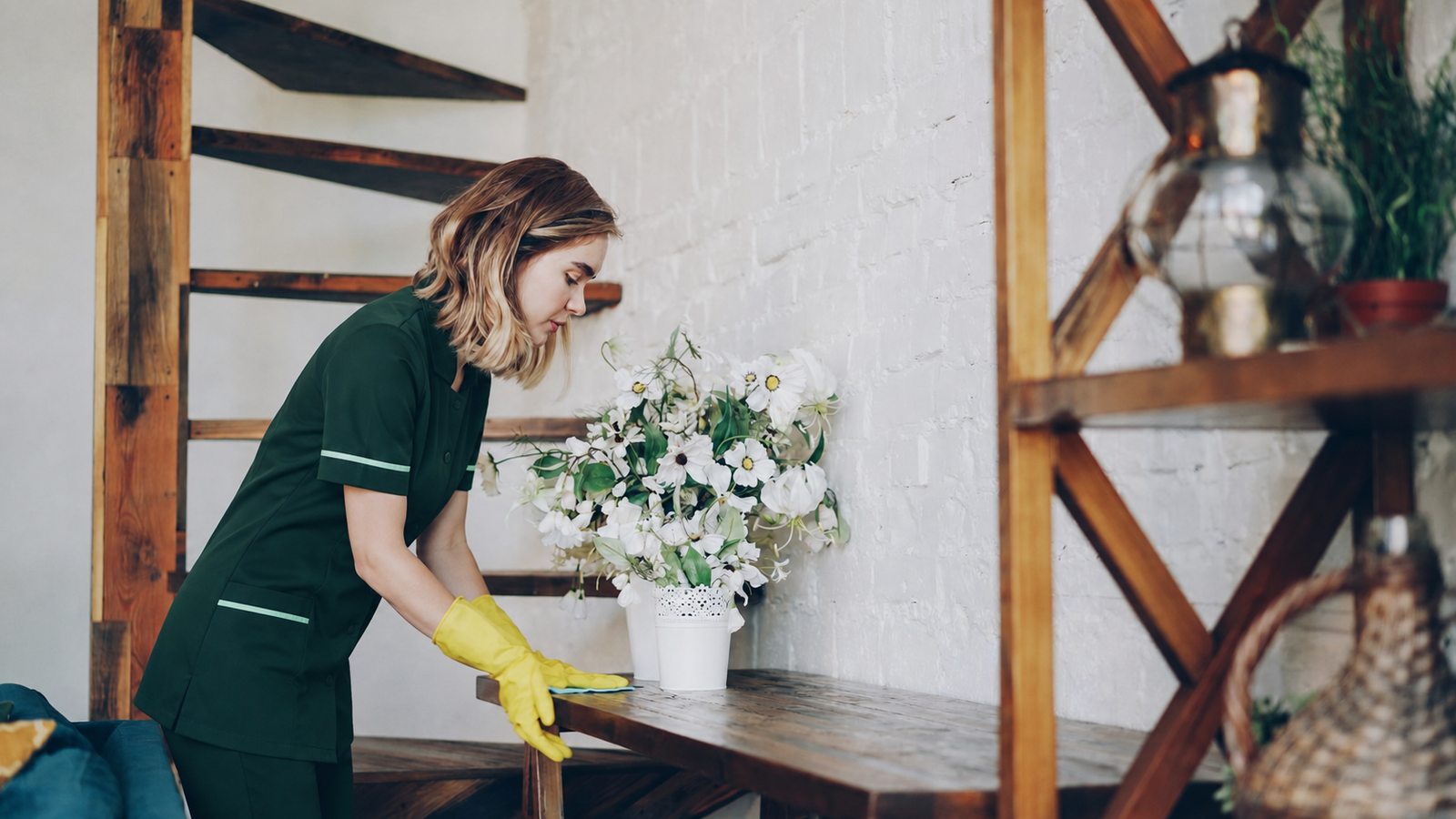 A blonde professional cleaner in a green uniform and yellow gloves carefully wiping a shelf